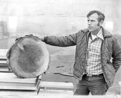 Man standing next to redwood log in black and white photo 