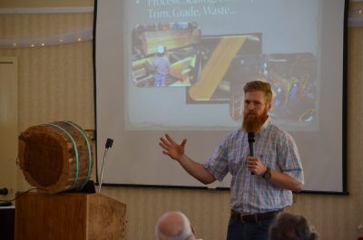 Man talking in microphone with a redwood round on the podium and a slide show in the background