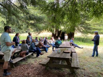 People at picnic tables listening to a presentation in a rustic setting