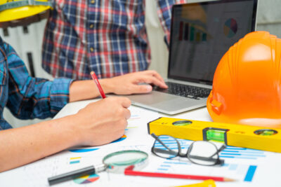 A desk with a hardhat, glasses a level and a person's arm taking notes