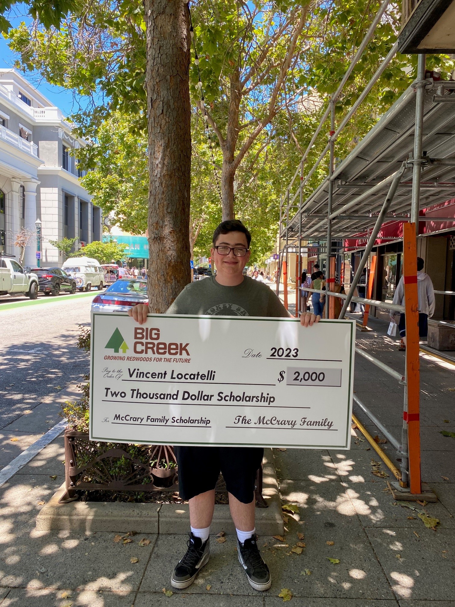 Young man holding big check in downtown santa cruz with trees in the background